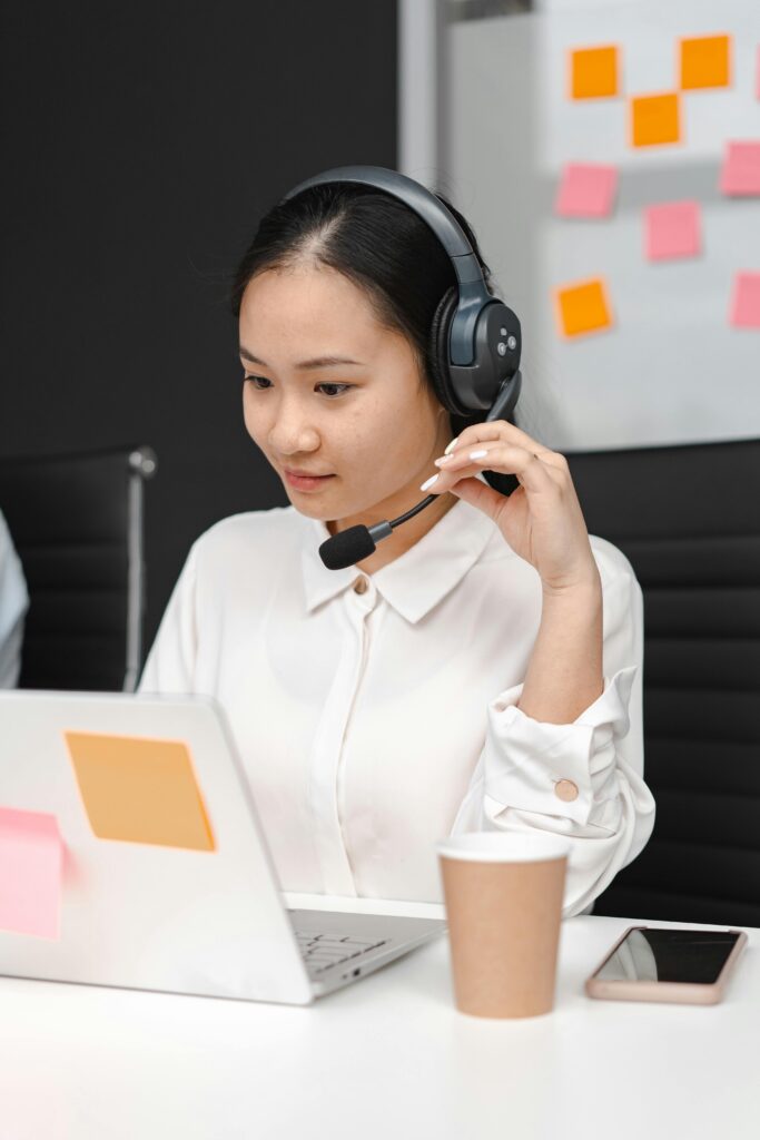 Asian woman using a laptop and headset for customer support in an office environment.